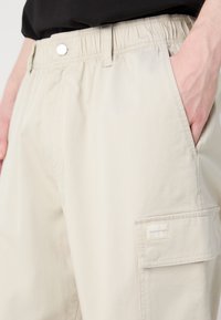 Világos bézs cargo short, könnyű anyagból készült, elasztikus derékkal, oldalsó zsebekkel és márkázott patkolású zsebbel.