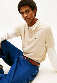 Beige Poloshirt mit weißen Akzenten, getragen über einem langen weißen Hemd. Dunkelblaue Jeans mit einem braunen Gürtel mit Logodetail.