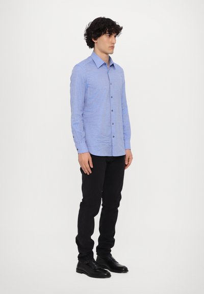 Camisa de manga larga a cuadros azul claro con cuello abotonado, combinada con pantalones ajustados negros y zapatos negros, de pie sobre un fondo blanco.