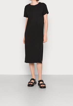 Person trägt ein schlichtes schwarzes Midikleid mit kurzen Ärmeln und seitlichen Schlitzen sowie schwarze Plateau-Sandalen mit Schnallen und steht auf einem grauen Boden.