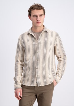 Mand iført en beige og gråstribet skjorte med knapper over en beige t-shirt og brune bukser, stående med den ene hånd i lommen.