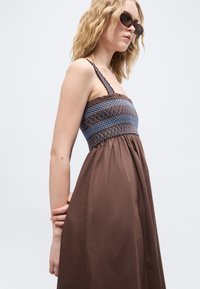 Γυναίκα που φοράει καφέ αμάνικο φόρεμα με μπλε σκούρο μοτίβο στο μπούστο και σκούρα γυαλιά ηλίου, ποζάροντας μπροστά σε απλό φωτεινό φόντο.