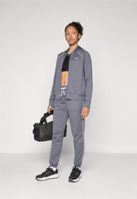 Grå treningsjakke med hette, matchende grå bukser, svart sports-BH og svarte joggesko. Har hvite detaljer og en svart treningsbag.