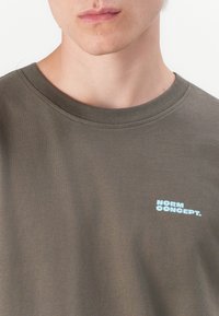 Olijfgroene katoenen t-shirt met een ronde hals, voorzien van een lichtblauwe "NORM CONCEPT." logo op de linkerborst. Soepele textuur.