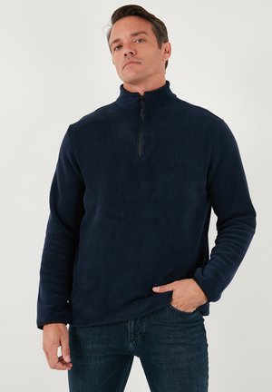 Mornarsko plava flis majica s polu-zip ovratnikom, dugim rukavima i mekanom teksturom. Ima suptilan sjaj i strukturni kroj.