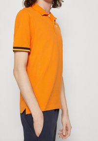 Personne portant un polo à manches courtes orange vif avec des poignets rayés noirs et jaunes, se tenant avec une main dans la poche de son pantalon marine.