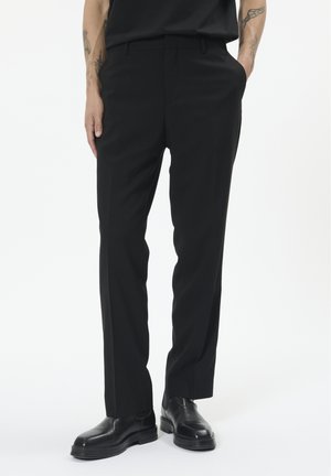 Pantalon noir avec une coupe ajustée, devant plat, poches latérales et une texture de tissu lisse. Porté avec des chaussures noires à bouts ronds.