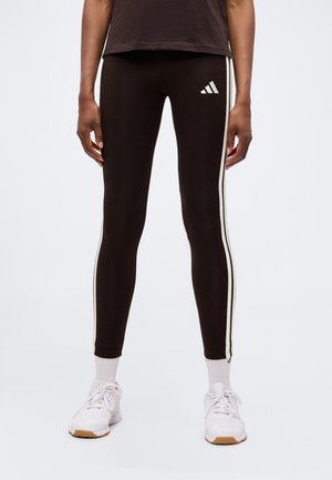 Personne portant des leggings noirs Adidas avec des bandes blanches sur les côtés, des chaussettes blanches et des chaussures de sport blanches, debout devant un fond uni.