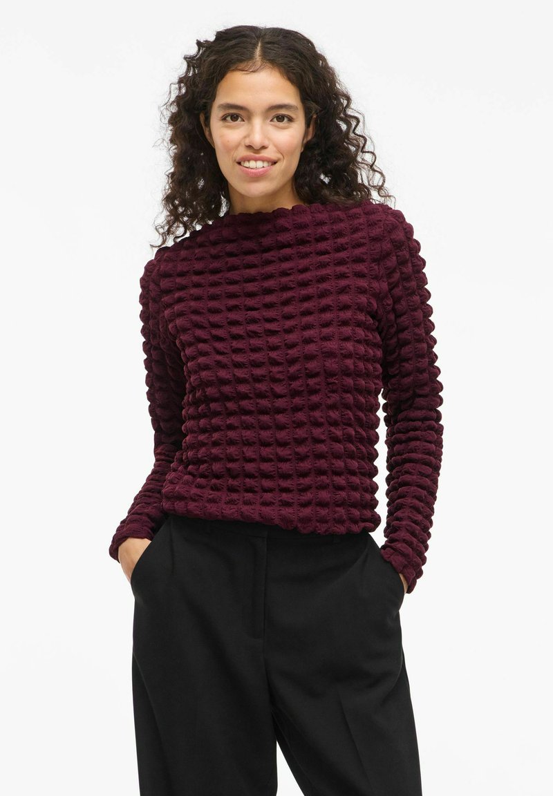 Bordeaux tekstureret sweater med et rutenetdesign, der har lange ærmer og høj hals; parret med løse sorte bukser og hænderne i lommerne.