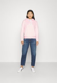 Felpa rosa chiaro con cappuccio e coulisse, dotata di tasca frontale, con il logo di Alpha Industries. Abbinata a jeans blu e sneakers bianche.