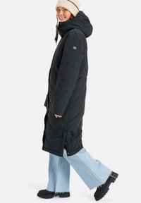 Abrigo largo negro acolchado con capucha, con diseño acolchado diagonal, aberturas laterales, combinado con vaqueros de pierna ancha azul claro y botas negras.