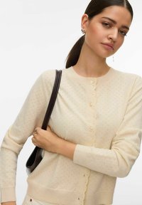 Cardigan tricoté de couleur crème avec une patte de boutonnage festonnée, présentant de petits motifs ajourés et des poignets côtelés. Porté avec un sac d'épaule sombre.