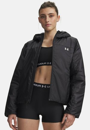 Vrouw met een zwart Under Armour jack met capuchon, een zwarte sportbeha en zwarte shorts, staand tegen een effen witte achtergrond.