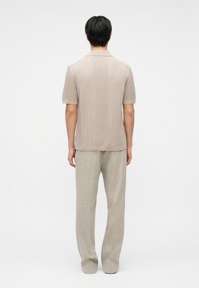 Άνδρας που στέκεται με την πλάτη γυρισμένη, φοράει μπεζ πλεκτό πόλο με κοντά μανίκια και χαλαρά, ανοιχτόχρωμα παντελόνια, σε απλό φόντο.