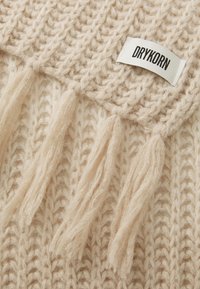 Écharpe en tricot beige avec des franges et une étiquette en tissu portant l'inscription "DRYKORN." Texture douce et motif en relief visibles en gros plan.
