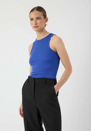 Débardeur bleu côtelé avec un col montant et des grandes emmanchures, associé à un pantalon noir ajusté avec poches et une texture lisse.