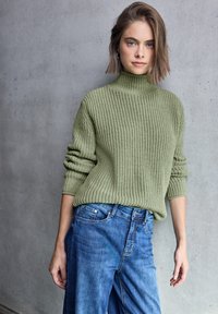 Groen geribbeld breiselsweater met hoge hals, gecombineerd met blauwe wijde denim jeans, weergegeven tegen een neutrale betonnen achtergrond.