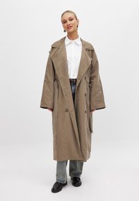 Svetlosandrový trenčkot s voľným strihom, širokým golierom, dvojradovým zapínaním a veľkými prednými vreckami, doplnený o pohodlné modré džínsy.