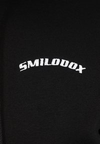 Tissu noir avec une texture côtelée présentant un texte brodé blanc indiquant "SMILODOX" dans un arc courbé. Design simple et minimal.