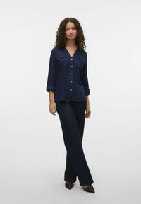Blusa blu navy con bottoni, due tasche sul petto, maniche lunghe arrotolate ai polsini, tessuto leggero, abbinata a pantaloni in denim scuro e scarpe a punta.