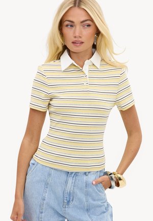 Femme blonde portant un polo rayé jaune à manches courtes avec un col blanc et un jean bleu clair, main dans la poche et bracelets dorés au poignet.