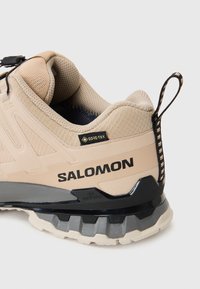 Baskets de sport beiges avec une surface texturée, étiquette GORE-TEX, accents noirs et semelle rugueuse à motifs. Dotées d'une languette à l'arrière.
