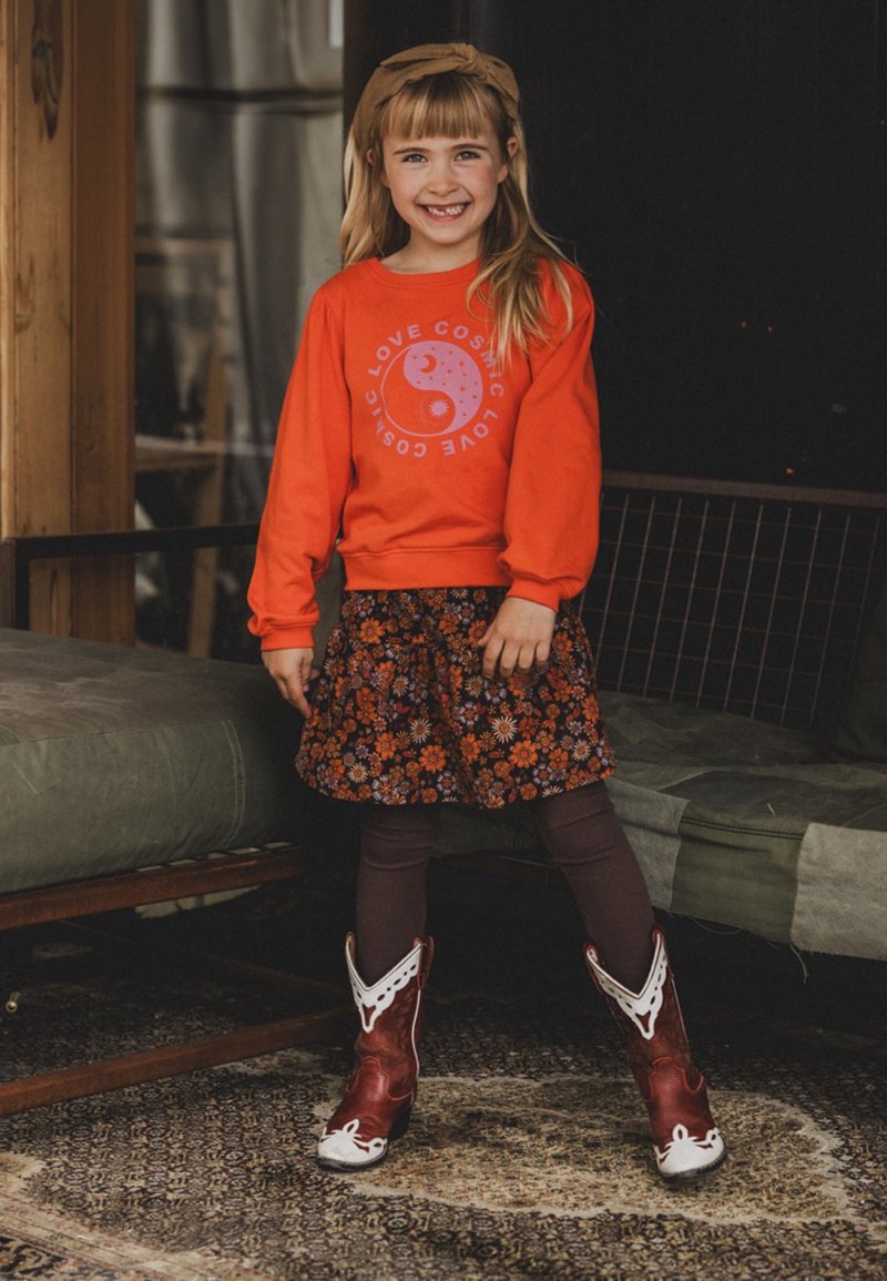 Rood sweatshirt met "Love Cosmic" graphic, bloemenpatroon rok, bruine legging, en bruine cowboy laarzen met witte accenten en stiksels.