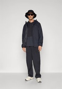 Juodas išorinis švarkas su gaubtu dėvimas virš tamsios trumpomis rankovėmis marškinėlių, derinamas su juodomis krovinių kelnėmis ir baltais sportbačiais. Aksesuarai: akiniai nuo saulės ir kibiro formos skrybėlė.