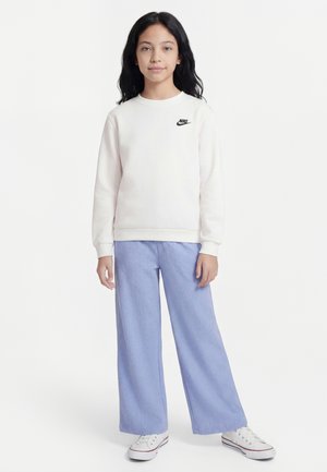 Dziewczyna z długimi czarnymi włosami, ubrana w biały sweter Nike, jasnoniebieskie spodnie o szerokich nogawkach oraz białe tenisówki, stoi zwrócona przodem.