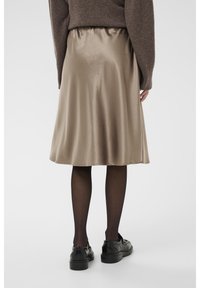 Jupe A-line en satin de couleur beige atténué, avec une texture douce et un ourlet légèrement évasé. Portée avec des collants noirs et des chaussures.