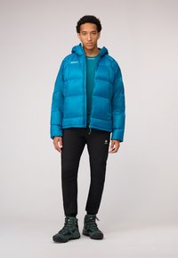 Teal puffer jacket with a hood, zippered front, and two pockets. Worn over a blue base layer, paired with black pants and green hiking boots.
