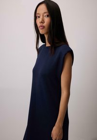 Ženska z dolgimi temnimi lasmi, oblečena v brezrokavno mornarsko modro obleko, stoji ob enotni svetlo sivi ozadju.