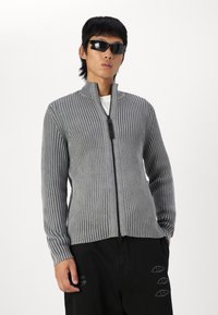 Suéter gris acanalado con cremallera y cuello alto, con detalle de cremallera negra, combinado con pantalones negros que presentan motivos de ojos. Modelo usando gafas de sol.
