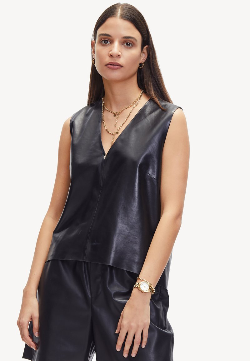 Schwarzes, ärmelloses Oberteil aus Leder mit V-Ausschnitt, ausgestattet mit einem Reißverschluss vorne und einer glatten Textur. Kombiniert mit schwarzen Lederhosen und goldener Schmuck.