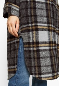 Cappotto a quadri in grigio, nero e senape con una texture morbida. Presenta un orlo curvo e una fessura laterale, indossato su jeans blu.