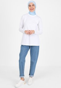 Weißes Langarmoberteil kombiniert mit einem hellblauen Hijab, hochgekrempelten hellblauen Jeans und weißen Sneakers mit Pastellakzenten. Einfaches und lässiges Design.