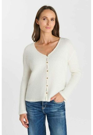 Donna che indossa un cardigan bianco peloso con bottoni e scollo a V e jeans blu, in piedi con una mano in tasca su uno sfondo semplice.