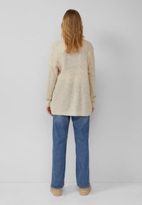 Beige Strickpullover mit relaxed Fit, der ein strukturiertes Muster, lange Ärmel und einen leicht verlängerten Rücken aufweist. Kombiniert mit blauen Denim-Jeans.