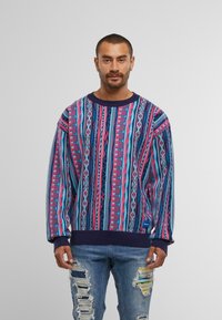 Mehrfarbiger Pullover mit geometrischen Mustern in Rosa, Blau und Lila. Verfügt über einen gerippten navyfarbenen Kragen und Saum, kombiniert mit zerrissenen Jeans.