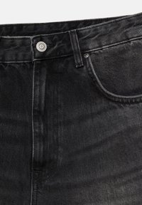 Gros plan sur un jean en denim noir montrant la ceinture, le bouton en métal, la boucle de ceinture, et les coutures de la poche avant.