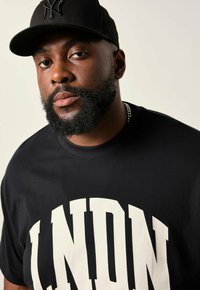Sort bomulds T-shirt med stort hvidt "LNDN" tryk. Har rund halsudskæring og korte ærmer, sammen med en sort baseballkasket.