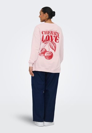 Nainen, jolla on tummat hiukset nutturalla, hänellä on vaaleanpunainen collegepaita, jossa on punaisia kirsikoita ja teksti "Cherry Love", tummansiniset housut ja valkoiset kengät, hän katsoo pois päin.
