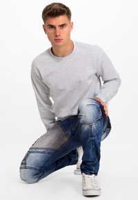 Lichtgrijze sweatshirt met lange mouwen, gecombineerd met blauwe denim jeans met een vervaagd, patchworkontwerp en witte sneakers.