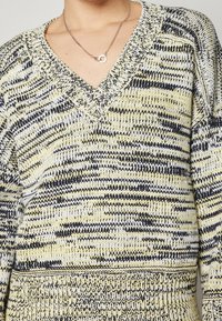 Eine Person trägt einen gelb-, schwarz- und weiß gemusterten V-Ausschnitt-Pullover und eine Silberkette mit einem runden Verschluss.