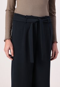 Navy wide-leg trousers with a tied waistband, featuring pleats and a smooth texture. Worn with a ribbed, light brown long-sleeve top.