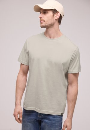 Mand iført beige kasket, lysegrå t-shirt med korte ærmer og blå jeans, kigger til venstre mod en ensfarvet baggrund.