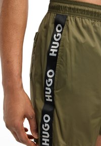 Õliverohelised sädeleva viimistlusega lühikesed püksid, millel on must küljeriba, millel on valge tekst "HUGO". Elastne vöökoht ja küljetasku detail.