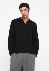 Giovane uomo con capelli corti scuri, che indossa un maglione nero a maniche lunghe e pantaloni grigi plissettati, in piedi con le mani in tasca su uno sfondo bianco.