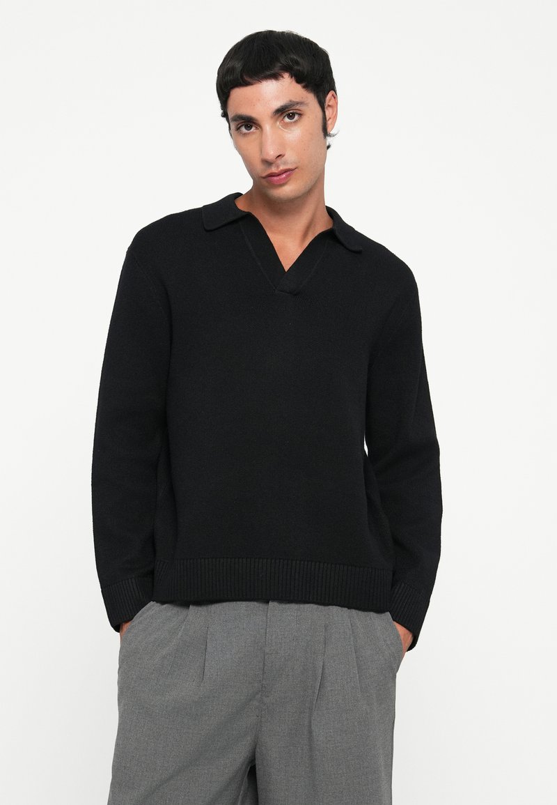 Giovane uomo con capelli corti scuri, che indossa un maglione nero a maniche lunghe e pantaloni grigi plissettati, in piedi con le mani in tasca su uno sfondo bianco.