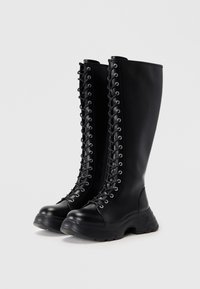 Ψηλές μαύρες δερμάτινες μπότες με χοντρές σόλες και σχέδιο κορδονιών μπροστά σε λευκό φόντο.